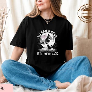 A woman wearing a black t-shirt with a graphic design and text, sitting on a bed with a vase of flowers in the background.