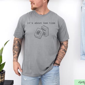 A man wearing a grey t-shirt with a graphic design and text printed on it, standing in front of a plant and a denim jacket.