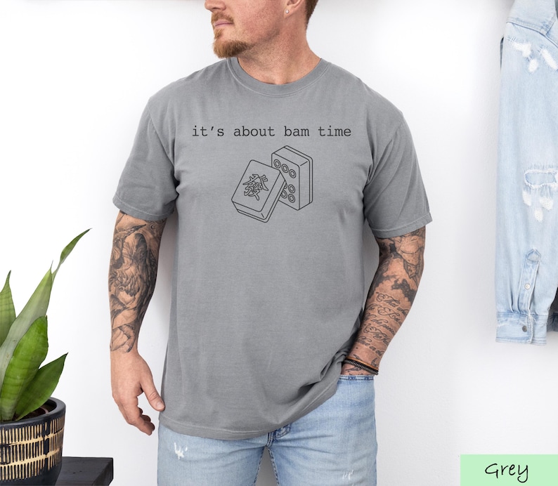 A man wearing a grey t-shirt with a graphic design and text printed on it, standing in front of a plant and a denim jacket.
