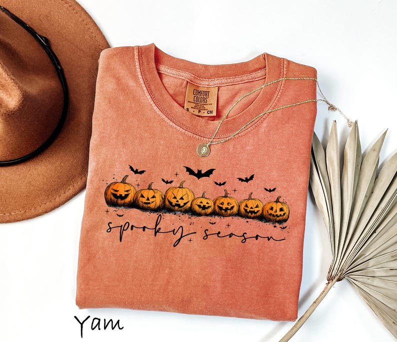 May include: A coral-colored t-shirt with a crew neck featuring a row of carved jack-o'-lanterns and black bats. The words "spooky season" are written in a script font. A gold necklace and a brown hat are also visible.