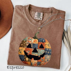 May include: Espresso-colored t-shirt with a patchwork pumpkin design. The pumpkin features a black carved face and is made of various fabric patterns in orange, blue, and yellow. A silver necklace with a pendant rests on the shirt.