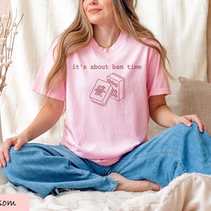 A woman wearing a pink t-shirt with a graphic design sits cross-legged on a bed, smiling.