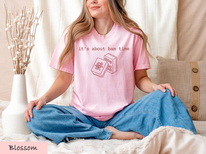 A woman wearing a pink t-shirt with a graphic design sits cross-legged on a bed, smiling.