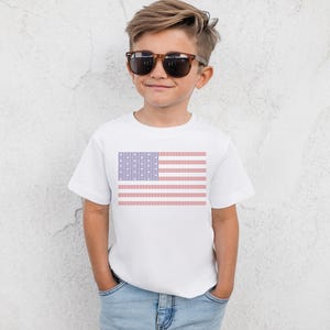 A young boy wearing a white t-shirt with an American flag design and sunglasses stands against a white wall.