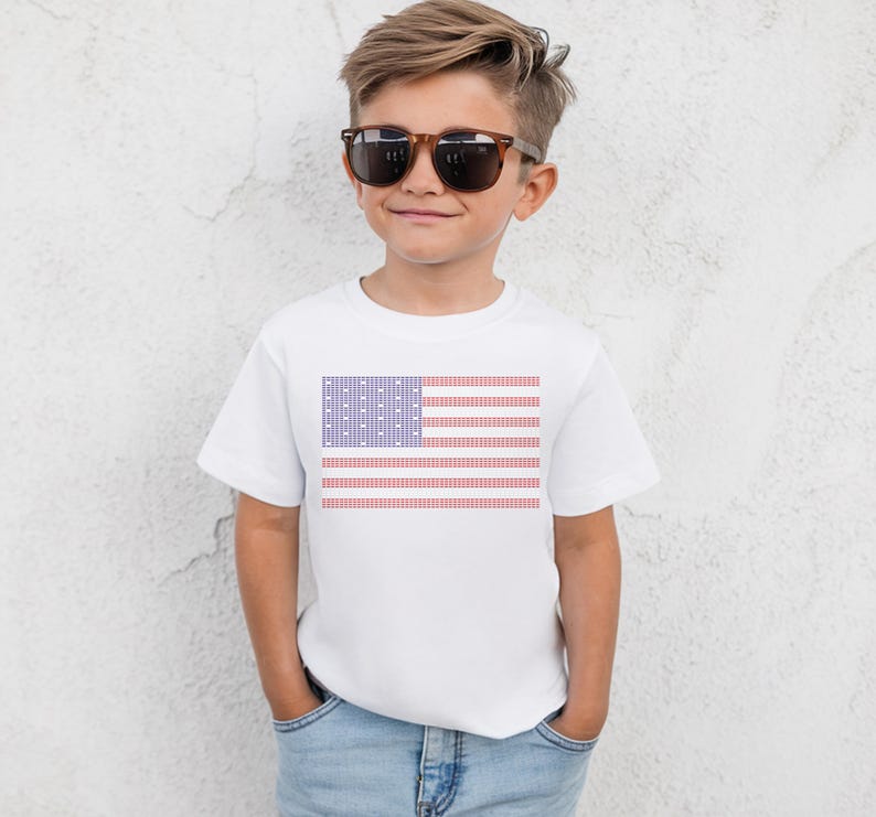 A young boy wearing a white t-shirt with an American flag design and sunglasses stands against a white wall.