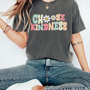 Puede incluir: Una camiseta gris antracita con un gr&aacute;fico de estilo retro que dice "Choose Kindness" con un dise&ntilde;o de margarita y cara sonriente.