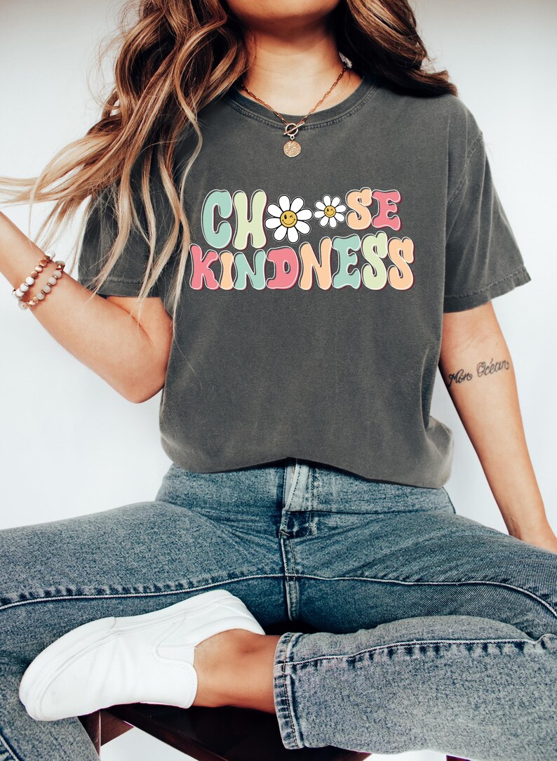 Puede incluir: Una camiseta gris antracita con un gr&aacute;fico de estilo retro que dice "Choose Kindness" con un dise&ntilde;o de margarita y cara sonriente.