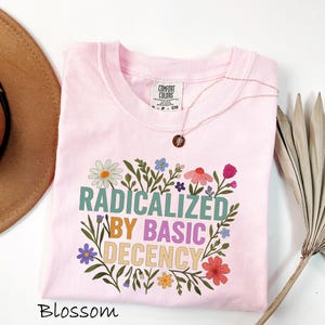 May include: A light pink t-shirt with the words "RADICALIZED BY BASIC DECENCY" in a floral design. The shirt is a Comfort Colors brand. A gold necklace with a pendant is on the shirt. The word "Blossom" is at the bottom.