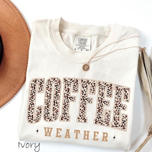 May include: An ivory-coloured t-shirt featuring the words "COFFEE WEATHER" in a brown leopard print design. The shirt has a crew neck and short sleeves. A gold necklace with a pendant is draped on the shirt.