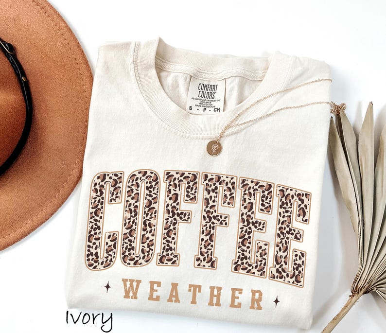 May include: An ivory-coloured t-shirt featuring the words "COFFEE WEATHER" in a brown leopard print design. The shirt has a crew neck and short sleeves. A gold necklace with a pendant is draped on the shirt.