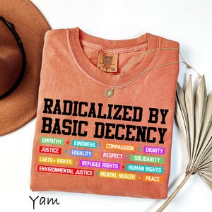 May include: A coral-colored t-shirt with the text "RADICALIZED BY BASIC DECENCY" in black block letters. Below, colorful words like "EMPATHY," "JUSTICE," and "LGBTQ+ RIGHTS" are displayed. A gold necklace and a brown hat are also visible.