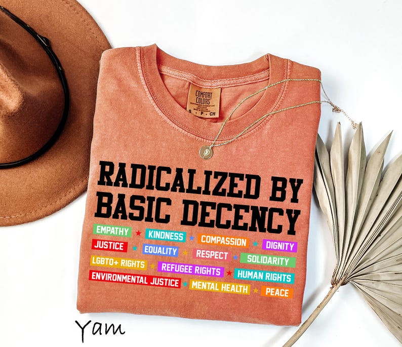 May include: A coral-colored t-shirt with the text "RADICALIZED BY BASIC DECENCY" in black block letters. Below, colorful words like "EMPATHY," "JUSTICE," and "LGBTQ+ RIGHTS" are displayed. A gold necklace and a brown hat are also visible.