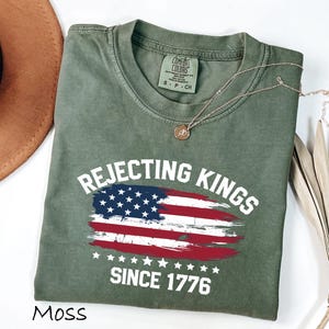 May include: Moss green t-shirt with the text "Rejecting Kings Since 1776" and an American flag design. The shirt is made of a soft material and has a crew neck. A gold necklace is draped across the shirt.