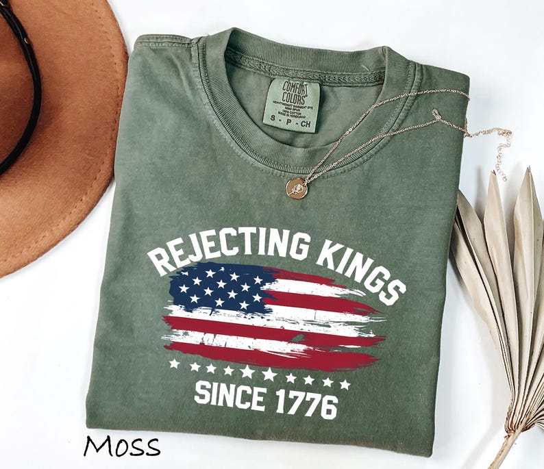 May include: Moss green t-shirt with the text "Rejecting Kings Since 1776" and an American flag design. The shirt is made of a soft material and has a crew neck. A gold necklace is draped across the shirt.