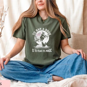A woman sitting cross-legged on a bed, wearing a green t-shirt with a graphic design and text.