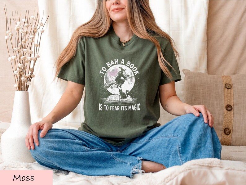 A woman sitting cross-legged on a bed, wearing a green t-shirt with a graphic design and text.