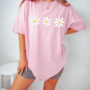 May include: A pink oversized t-shirt with a graphic of three white daisies with yellow centers.