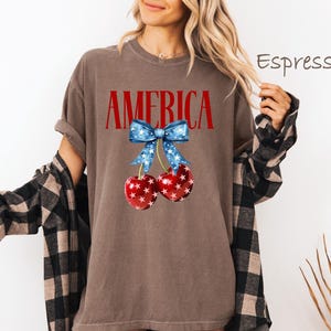 May include: A brown t-shirt with the word "AMERICA" printed in red letters. A red, white, and blue bow with stars is above two red cherries with white stars.