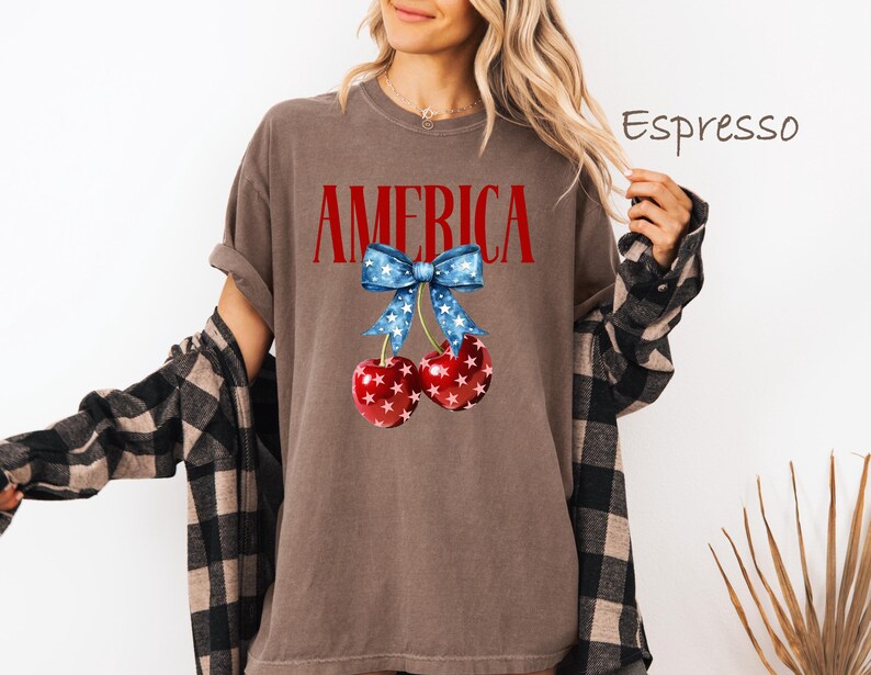 May include: A brown t-shirt with the word "AMERICA" printed in red letters. A red, white, and blue bow with stars is above two red cherries with white stars.