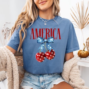 May include: A blue t-shirt with the word "AMERICA" printed in red letters. A red and white cherry with a blue and white star patterned bow is printed below the word.
