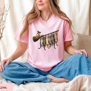 A woman wearing a pink t-shirt with a graphic of cats sitting on a broom is sitting cross-legged on a bed, smiling.