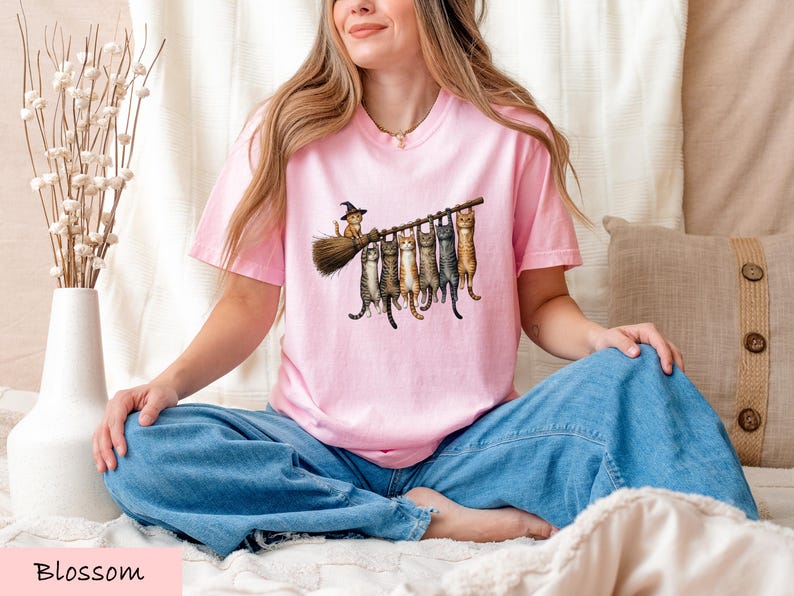 A woman wearing a pink t-shirt with a graphic of cats sitting on a broom is sitting cross-legged on a bed, smiling.