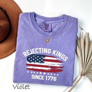 May include: A violet-colored t-shirt with the text "Rejecting Kings Since 1776" above a distressed American flag design. The shirt has a crew neck and short sleeves. A gold necklace and a brown hat are also in the image.