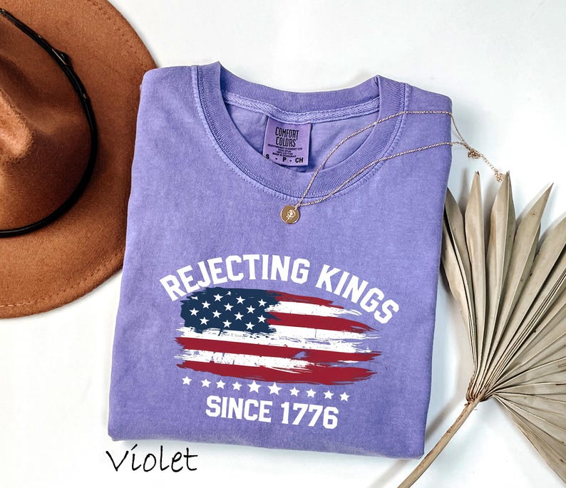 May include: A violet-colored t-shirt with the text "Rejecting Kings Since 1776" above a distressed American flag design. The shirt has a crew neck and short sleeves. A gold necklace and a brown hat are also in the image.