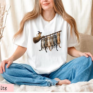 A woman wearing a white t-shirt with a graphic of cats sitting on a broom, smiling and sitting cross-legged on a bed.