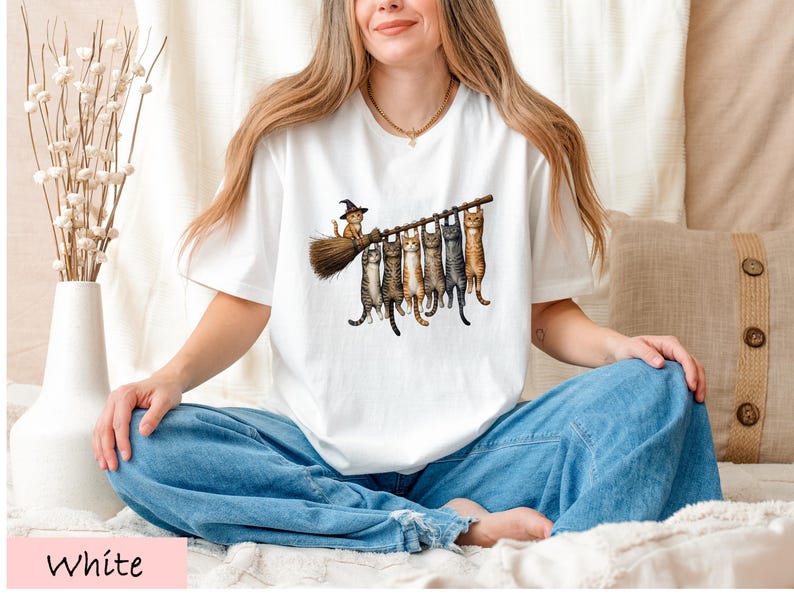 A woman wearing a white t-shirt with a graphic of cats sitting on a broom, smiling and sitting cross-legged on a bed.