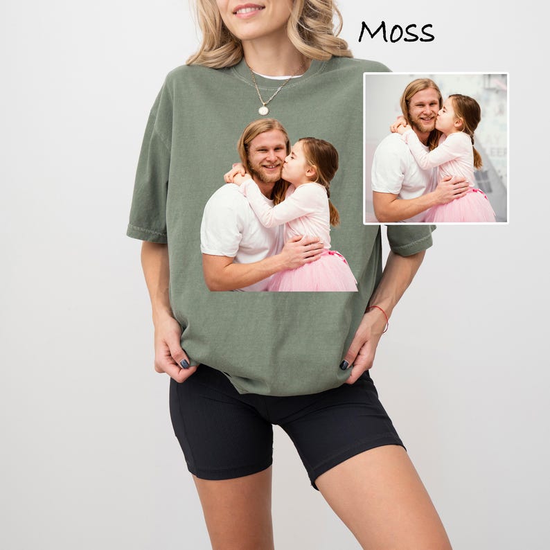 May include: A moss green t-shirt featuring a printed image of a man and a child. The image shows the man embracing the child, who is wearing a pink tutu. The word "Moss" is printed in black at the top right.