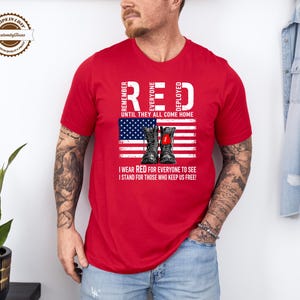 May include: Red t-shirt with the text "RED" and "REMEMBER EVERYONE DEPLOYED UNTIL THEY ALL COME HOME." The shirt features an American flag design with combat boots. The text "I WEAR RED FOR EVERYONE TO SEE, I STAND FOR THOSE WHO KEEP US FREE!" is also printed on the shirt.