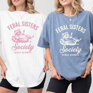 May include: A white t-shirt and a blue jeans colored t-shirt, both featuring a graphic of a raccoon and the text FERAL SISTERS Society NO RULES. NO REGRETS. The white shirt has pink text and graphics, while the blue shirt has white text and graphics.
