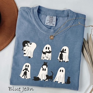 May include: Blue jean colored t-shirt featuring a playful design of black cats interacting with ghost figures. The shirt has a crew neck and short sleeves. A gold necklace with a pendant is draped across the shirt. The text "Blue Jean" is at the bottom.