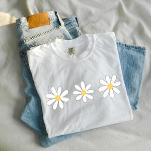 May include: White t-shirt with three yellow and white daisy flowers printed on the front. The shirt is folded and laid on top of a pair of blue jeans.