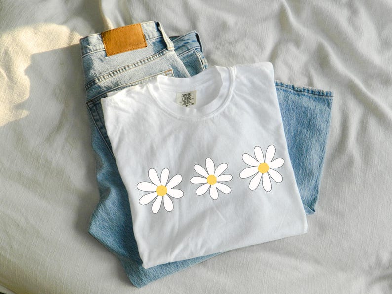 May include: White t-shirt with three yellow and white daisy flowers printed on the front. The shirt is folded and laid on top of a pair of blue jeans.