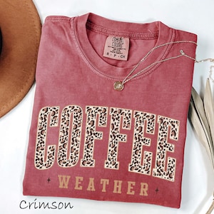 May include: Crimson-coloured t-shirt featuring the words "COFFEE WEATHER" in a leopard print design. The shirt has a crew neck and short sleeves. A gold necklace with a pendant is draped across the shirt.