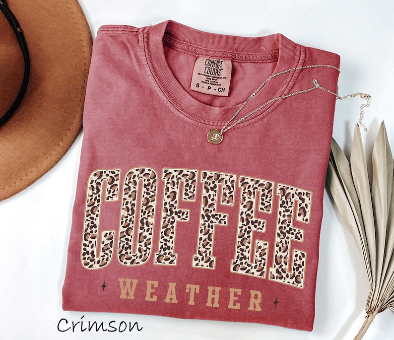 May include: Crimson-coloured t-shirt featuring the words "COFFEE WEATHER" in a leopard print design. The shirt has a crew neck and short sleeves. A gold necklace with a pendant is draped across the shirt.