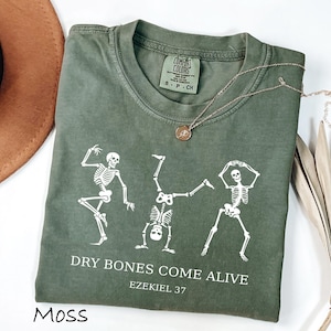 Peut inclure: T-shirt vert mousse à col rond et manches courtes. Le t-shirt présente un motif blanc de trois squelettes dansant et le texte "DRY BONES COME ALIVE" et "EZEKIEL 37". Un collier en or avec un pendentif est drapé sur le t-shirt.