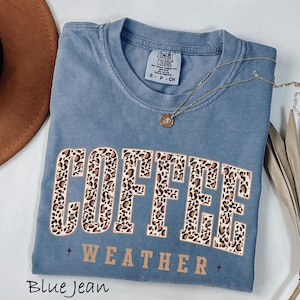 May include: Blue jean coloured t-shirt with the words "COFFEE WEATHER" in a leopard print design. The shirt has a crew neck and short sleeves. A gold necklace with a pendant is draped across the shirt.