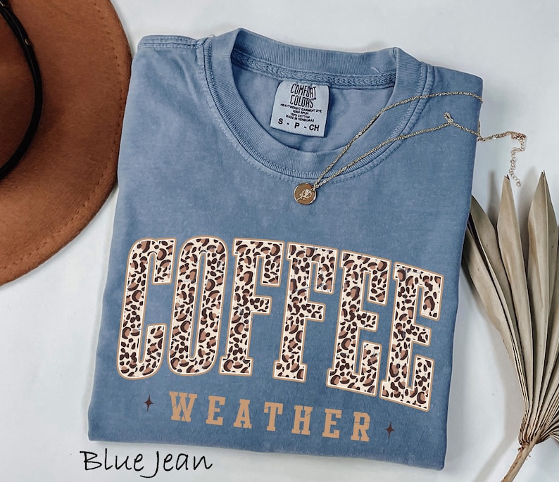May include: Blue jean coloured t-shirt with the words "COFFEE WEATHER" in a leopard print design. The shirt has a crew neck and short sleeves. A gold necklace with a pendant is draped across the shirt.