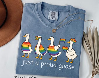 Puede incluir: Camiseta azul vaquero con un gráfico de cuatro gansos de dibujos animados en colores arcoíris, cada uno con un accesorio diferente. El texto "just a proud goose" está impreso debajo. Un collar dorado está drapeado sobre la camisa.