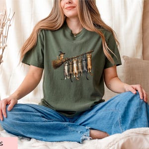 A woman sitting cross-legged on a bed, wearing a green t-shirt with a graphic of cats and a witch on a broomstick.