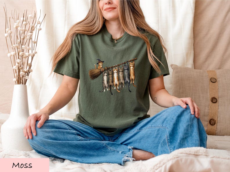 A woman sitting cross-legged on a bed, wearing a green t-shirt with a graphic of cats and a witch on a broomstick.