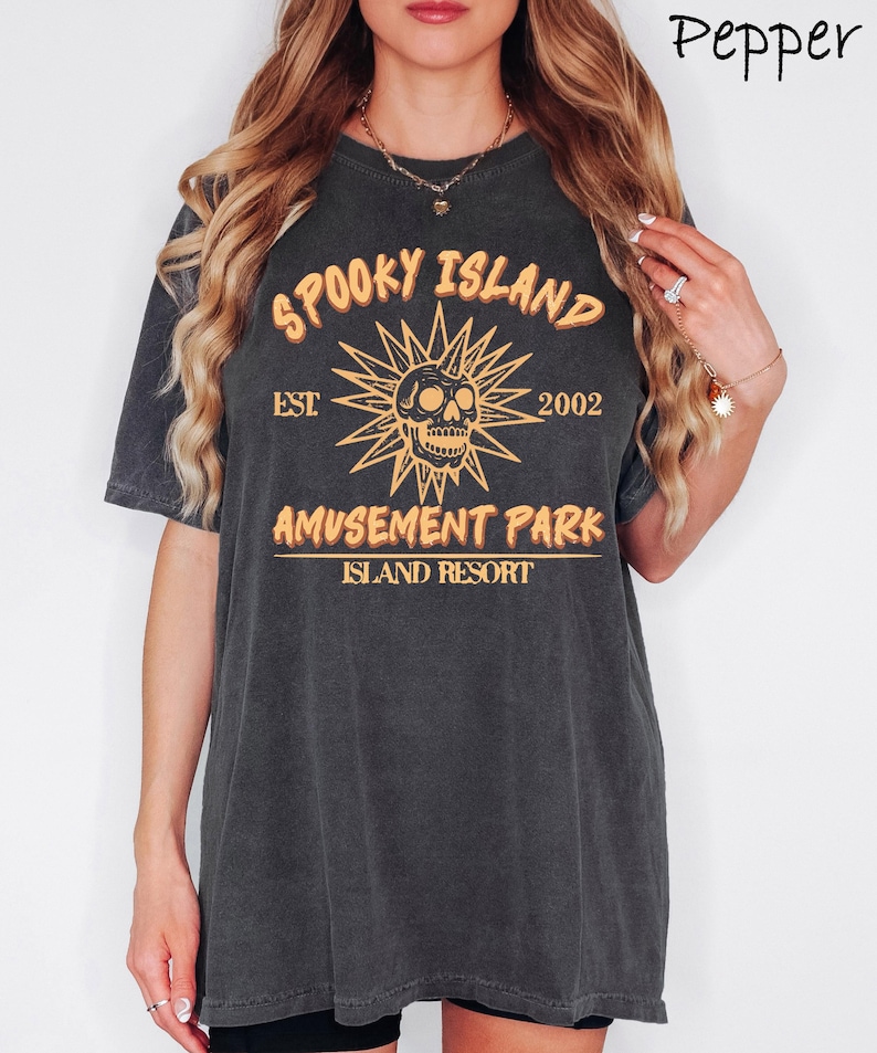 May include: Dark gray t-shirt with a distressed graphic. The design features the text "Spooky Island Amusement Park Island Resort" with a skull and sunburst graphic in orange. The shirt has short sleeves and a crew neck.