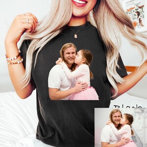 May include: Black t-shirt featuring a printed image of a man and a young girl. The image shows the man embracing the girl, who is kissing his cheek. The girl is wearing a pink tutu.