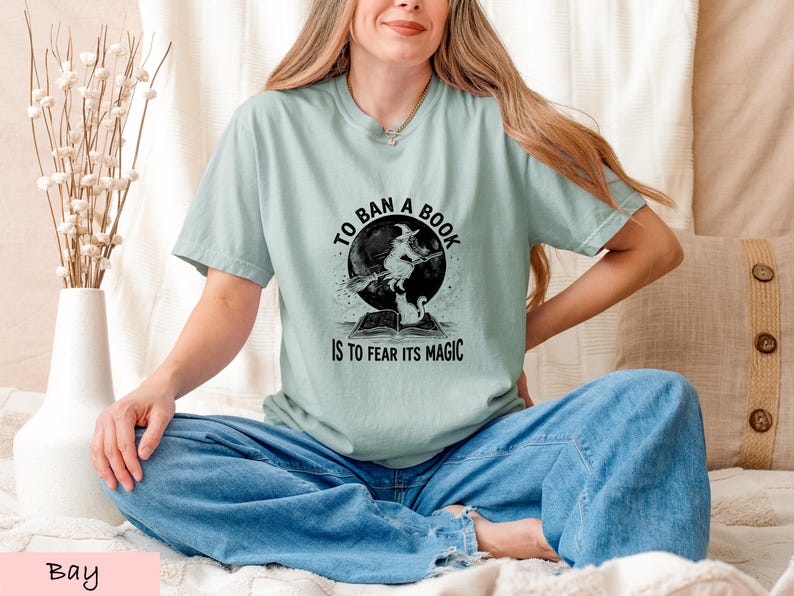 A woman wearing a light green t-shirt with a graphic design and text, sitting on a bed with a vase of flowers and a pillow nearby.