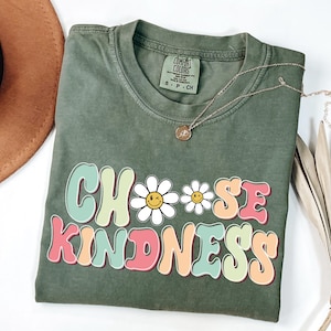 Puede incluir: Camiseta verde con el texto "Choose Kindness" en letras retro de colores con dos margaritas blancas con caras sonrientes.