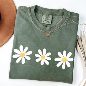 May include: A green t-shirt with a row of three white daisies with yellow centers. The shirt has the text "COMFORT COLORS" on the tag.