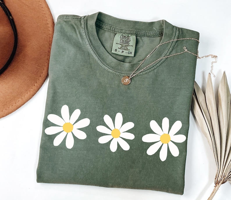 May include: A green t-shirt with a row of three white daisies with yellow centers. The shirt has the text "COMFORT COLORS" on the tag.